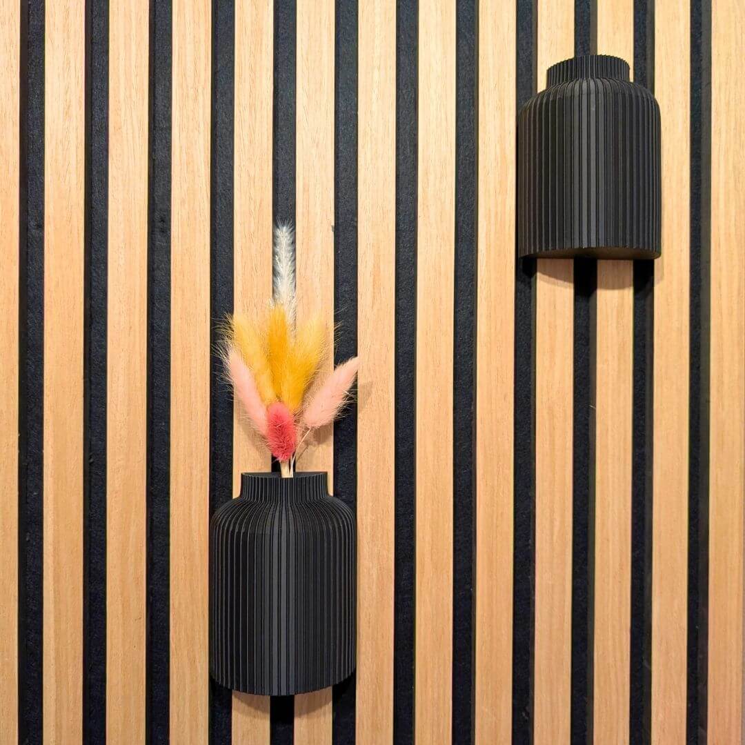 Zwei schwarze 3D-gedruckte Wandvasen auf einem Akustikpaneel: Eine puristisch leer und eine mit bunten Trockenblumen als moderne Wanddekoration ohne Bohren.